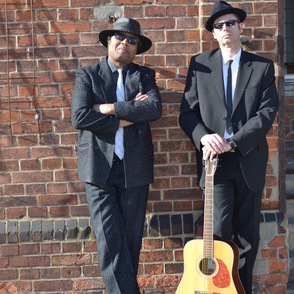 Foto des Duos in schwarzen Anzügen vor einer Ziegelwand mit Gitarre