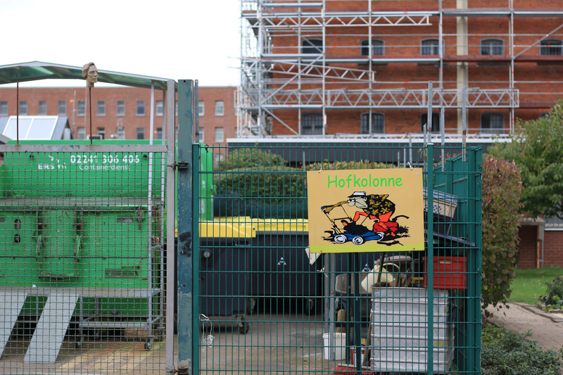 Das Schild "Hofkolonne" hängt am Zaun der den Bereich der Restmüllpresse abgrenzt. Im Hintergrund ist Haus 1 zu sehen.