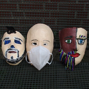 Drei handgefertigte bunt angemalte Masken liegen auf einem Gitter zum trocknen