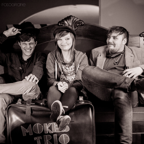 Foto des Trios auf einer Couch mit Füßen auf einem Koffer mit Bandlogo in SW